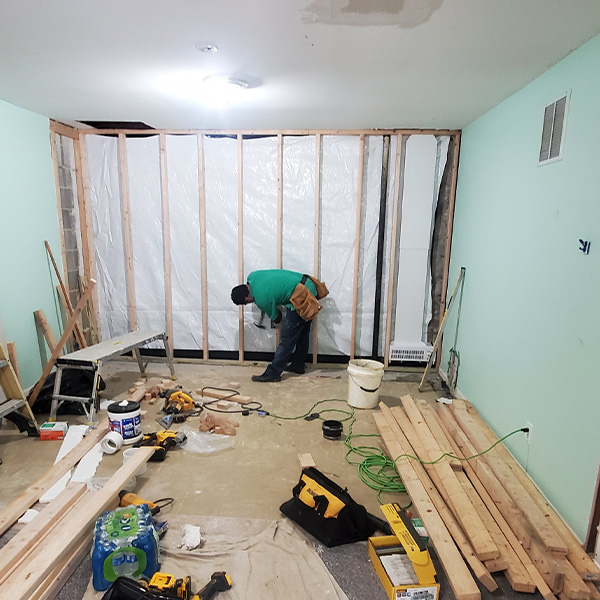 Worker in a green shirt and tool belt works on framing a wall in a room under construction